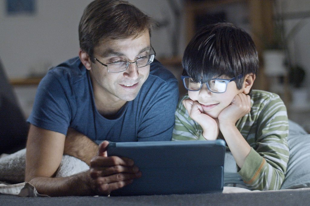 Photo and father and son looking at a tablet device together, their faces illuminated by it's glow.